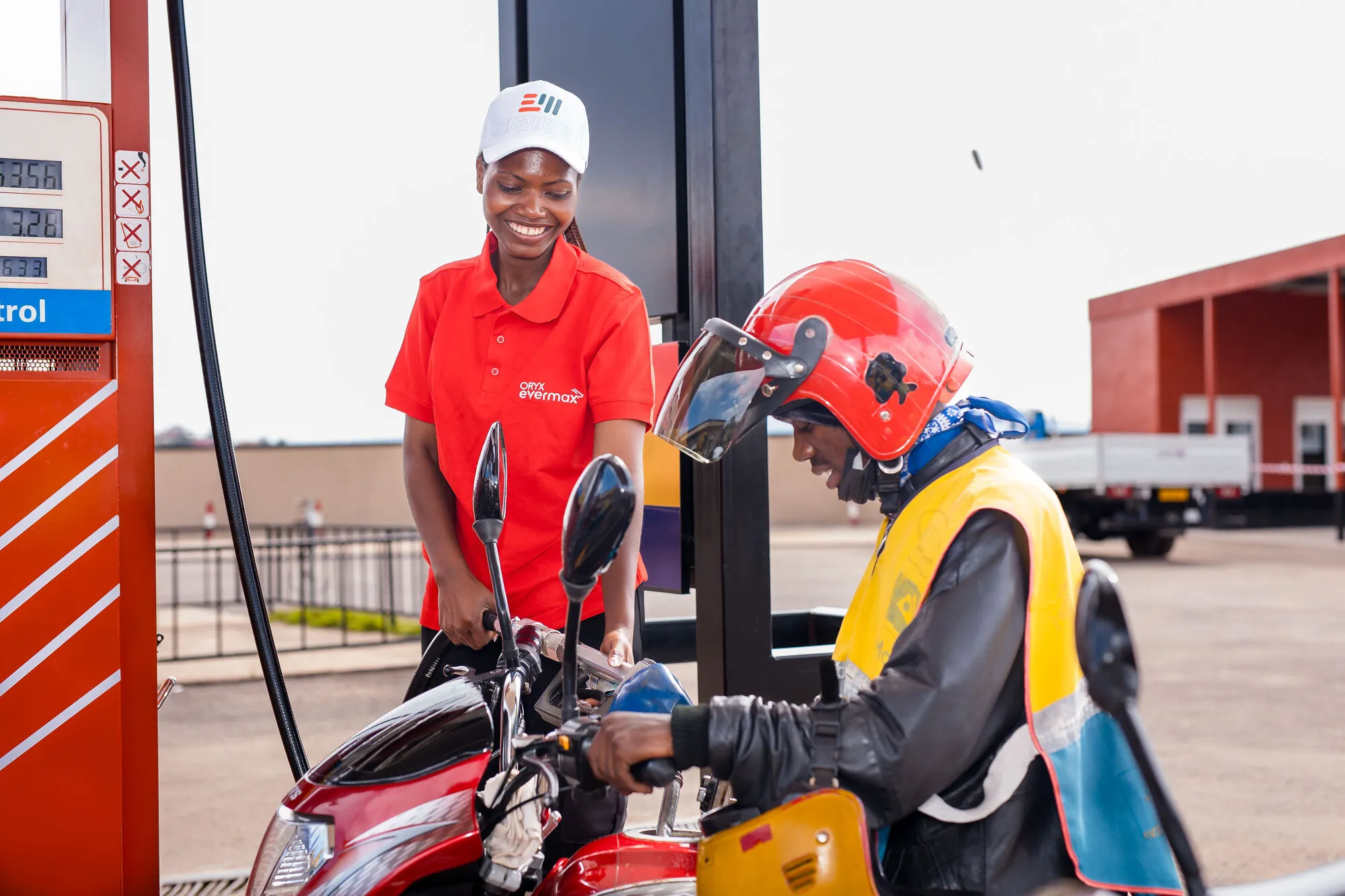 Attendant in red uniform fueling a motorcycle ridden by a person in a red helmet.